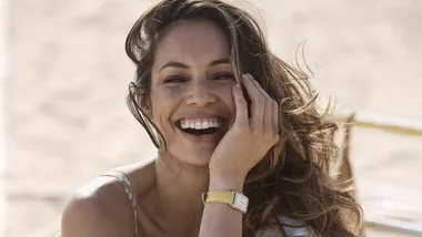 Smiling woman with long hair poses outdoors, hand on face, wearing a watch.
