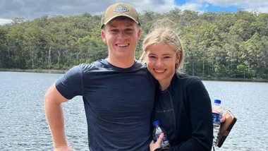 A smiling couple poses by a lake, surrounded by trees, holding water bottles on a cloudy day.