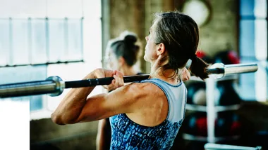 A woman in a gym lifting a barbell, focused and determined, wearing a blue patterned tank top.