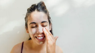 Woman applying face cream, smiling with eyes closed against a light background.