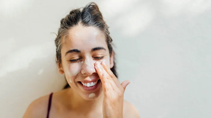 Woman applying face cream, smiling with eyes closed against a light background.
