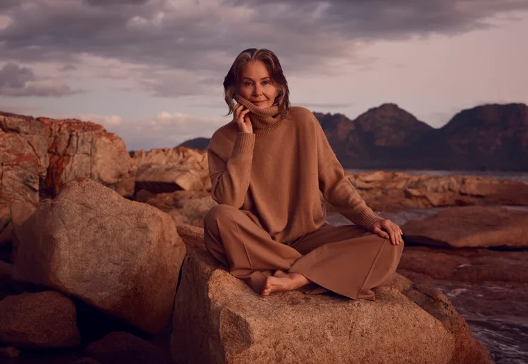 A woman in a brown sweater sits on rocks by the sea with mountains in the background, under a cloudy sky.