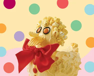 Whimsical butter sculpture of a bird with popcorn hair, chip beak, red bow, and polka dot background.