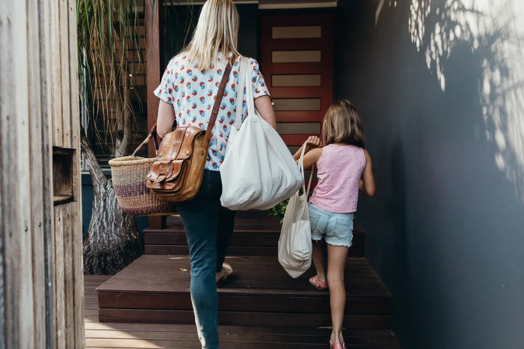 A woman and a child carrying bags walk up wooden steps toward a house with a red door.