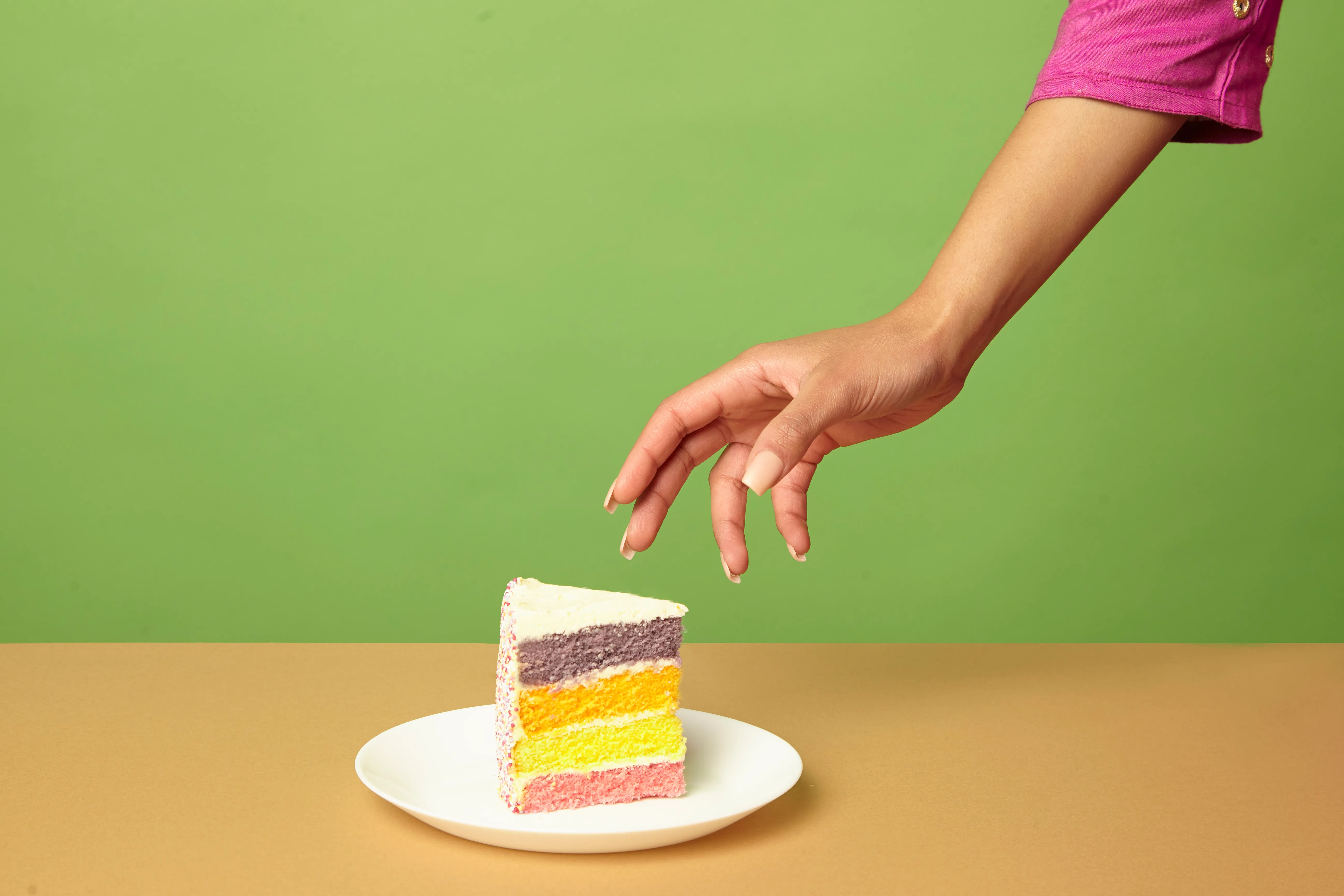 studio shot of woman's hand reaching for a slice of cake on a table
