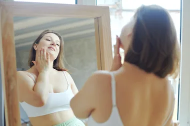 Woman in a white top looks at herself in the mirror, gently touching her face with her hand.
