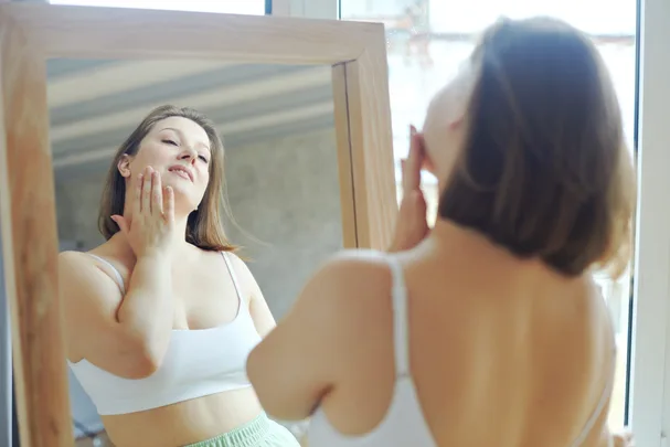 Woman in a white top looks at herself in the mirror, gently touching her face with her hand.