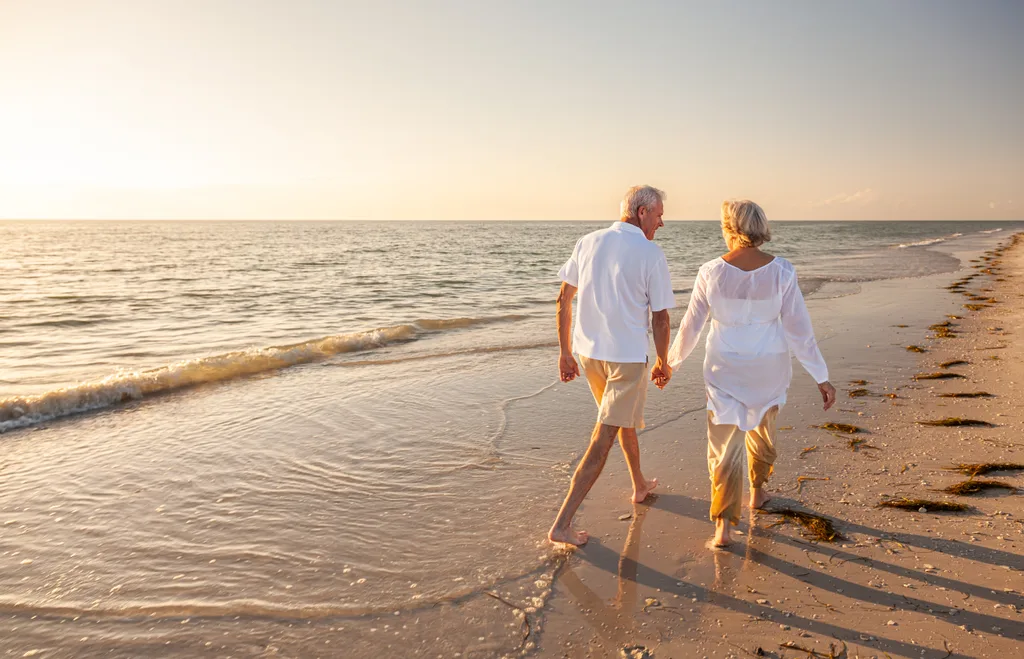 Happy senior man and woman old retired couple walking and holding hands on a beach at sunset.