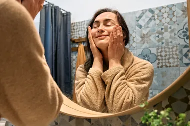 Person in a beige robe smiling while touching their face in a bathroom mirror.