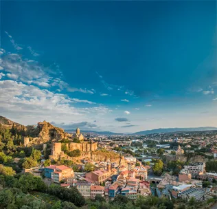 Panoramic view of Tbilisi, Georgia, featuring historic buildings, a fortress, and a sprawling cityscape under a blue sky.