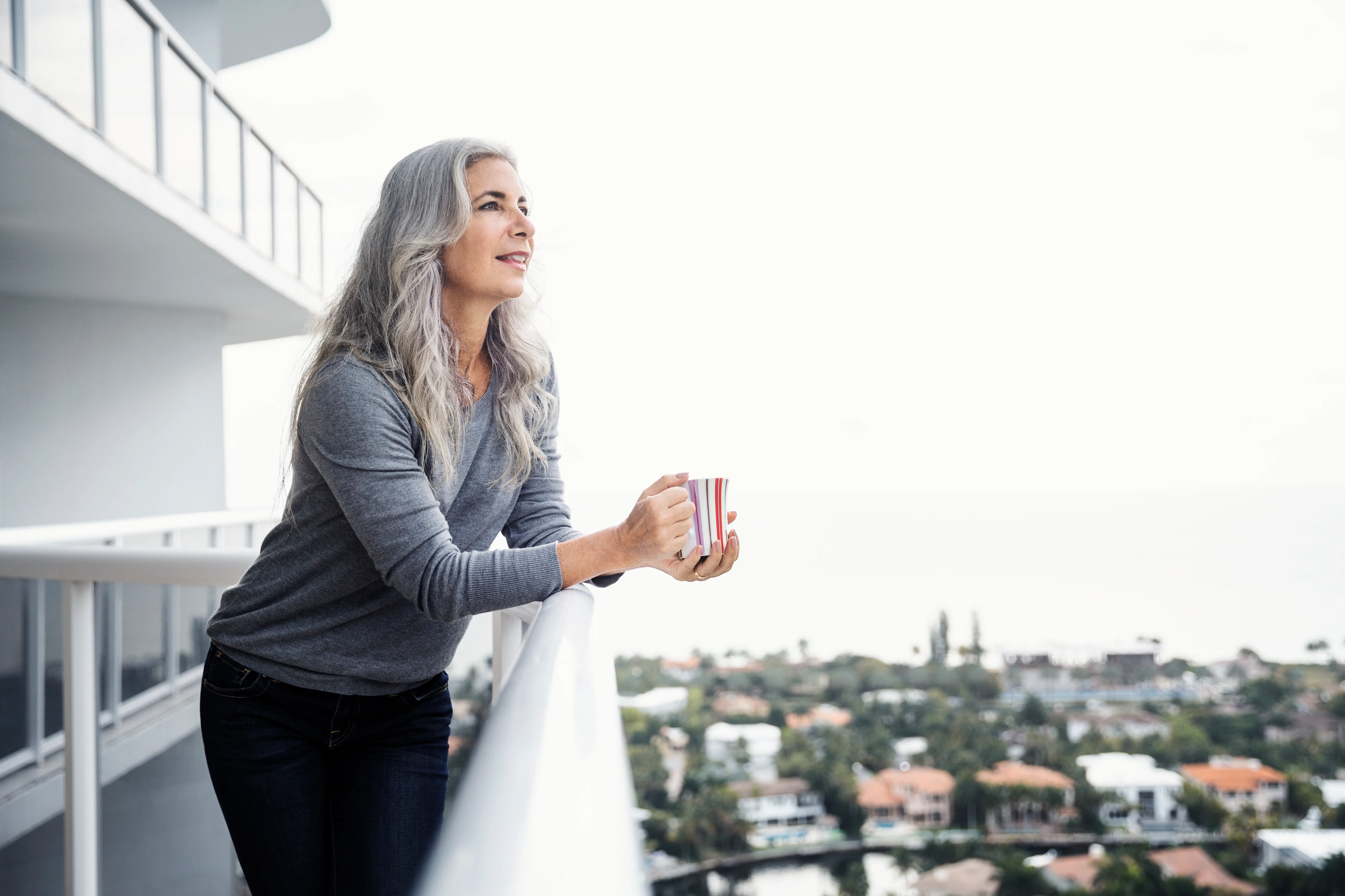 Grey haired woman drinking coffee looking over balcony.