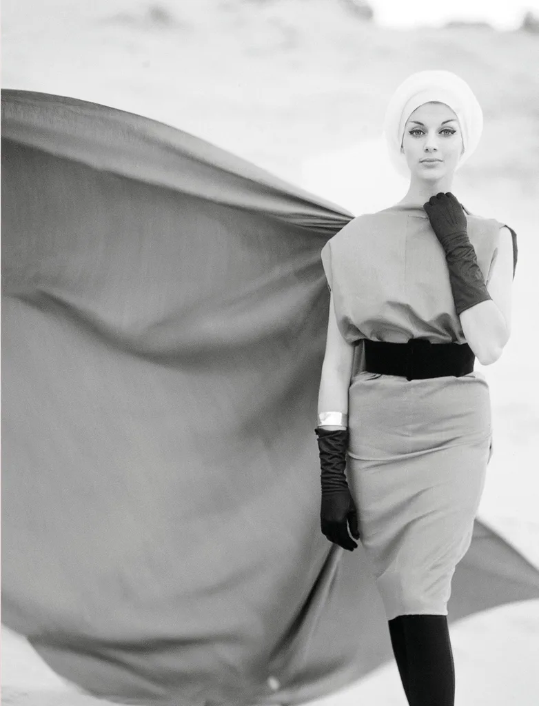 Elegantly dressed woman in black gloves and white turban, standing with a flowing cape in a desert setting.