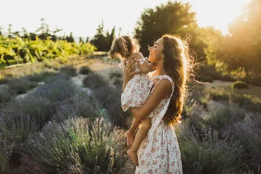 Mother holding child in a sunlit field, both wearing floral dresses, smiling joyfully in nature.