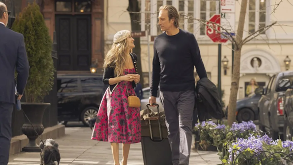 A man and a woman walking on a city sidewalk, with the man holding a suitcase and wearing casual attire.