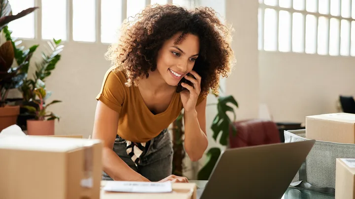 Woman smiles while talking on phone and using laptop, surrounded by boxes and plants in a well-lit room.