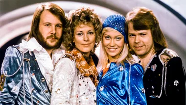 Four people in colorful, vintage costumes pose together, smiling, with a retro backdrop.