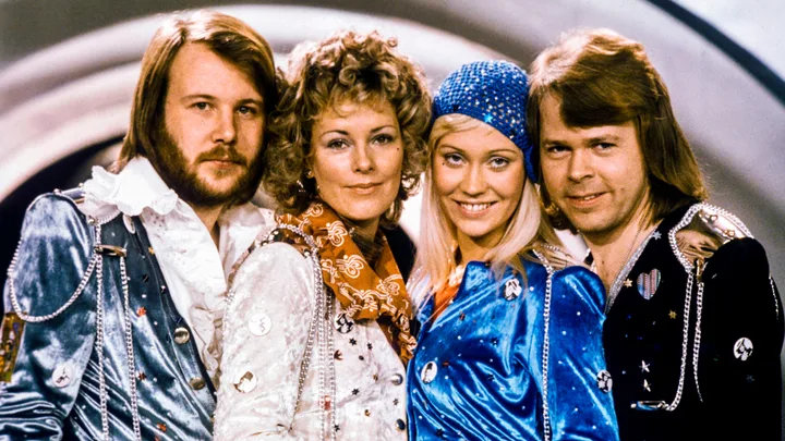 Four people in colorful, vintage costumes pose together, smiling, with a retro backdrop.