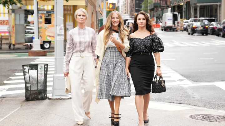 Three women walk confidently on a New York City street, stylishly dressed, carrying bags.