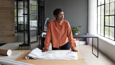 A woman in an orange blouse leans on a table with architectural plans in a modern, light-filled office space.