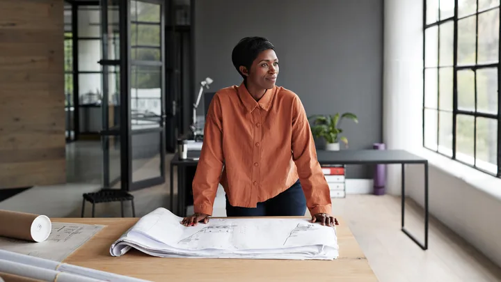 A woman in an orange blouse leans on a table with architectural plans in a modern, light-filled office space.