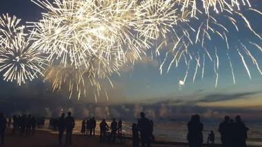 Silhouetted crowd watches bright fireworks display over a calm beach at dusk.