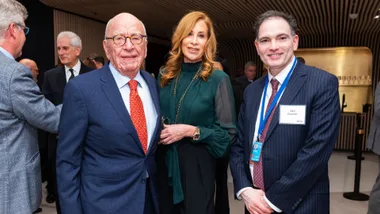 Three people in formal attire at an event, smiling. The man on the right wears a name badge reading "Dilip Chauhan."