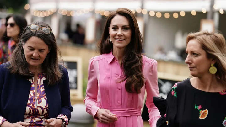 Three women walking outdoors, one in a pink dress smiling, others in patterned dresses, with blurred festive lights behind.