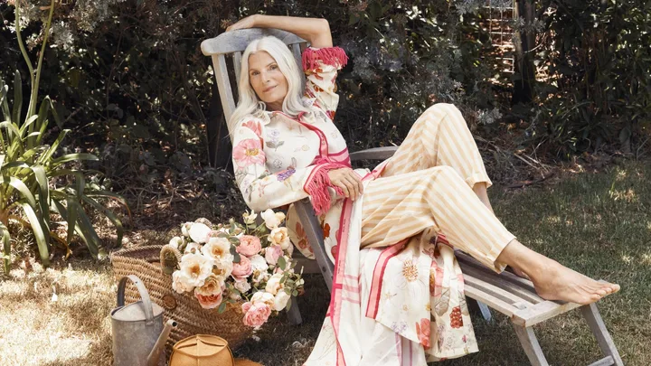 Woman with long gray hair lounging on a wooden chair in a garden, wearing floral outfit, with flowers nearby.