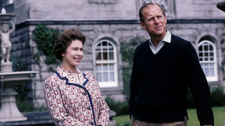 A woman smiles and stands beside a man outdoors, with a stone wall and greenery in the background.