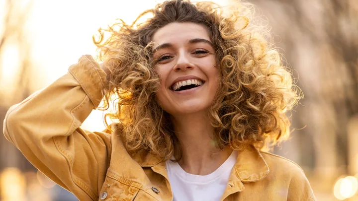 Smiling woman with curly hair in a yellow jacket under sunlight.