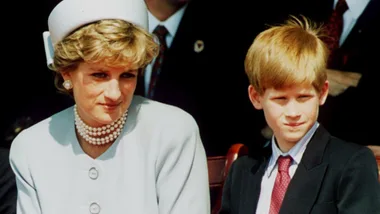 A woman in a white hat and pearls sits beside a boy in a suit and tie at an outdoor event.