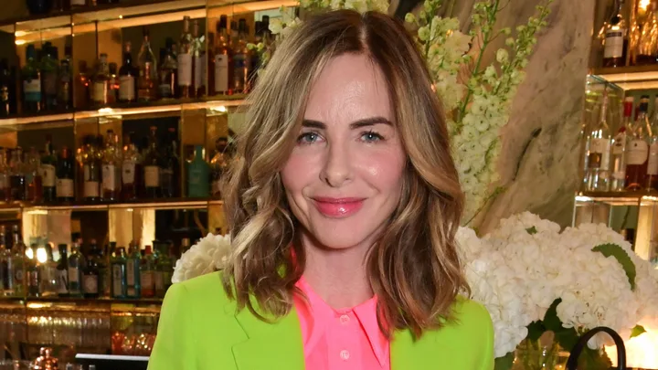 A woman with wavy hair in a bright green blazer stands in front of a bar with bottles and flowers.