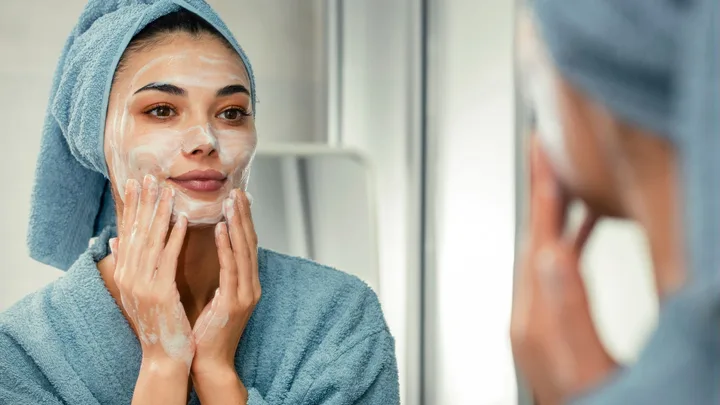 Woman in a blue towel applies facial cleanser while looking in the mirror.