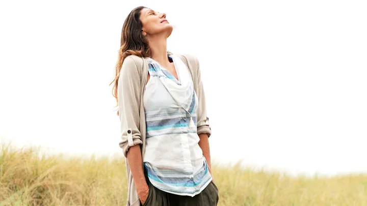 Woman enjoying nature, looking up with a smile, standing in a field with hands in pockets and wearing casual attire.