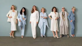 Seven women in stylish suits and dresses stand smiling against a light green background.