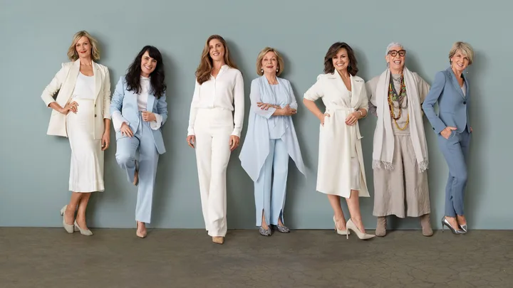 Seven women in stylish suits and dresses stand smiling against a light green background.