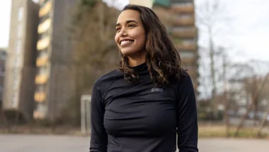 A woman in a black top smiles outdoors, with blurred buildings and trees in the background.