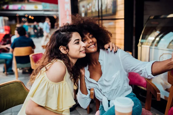 Two friends taking a selfie at an outdoor café, smiling and sitting close together.