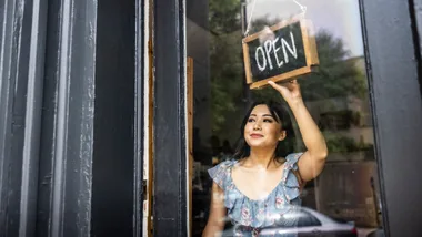 Woman holding up an open sign in front of shop to represent starting a small business