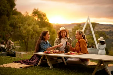 three friends drink wine at a picnic table while the sun sets