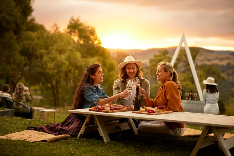 three friends drink wine at a picnic table while the sun sets