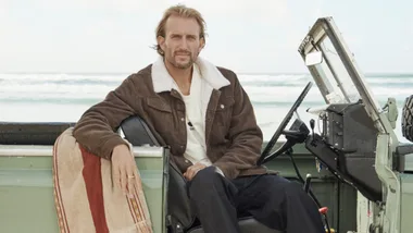 Man sitting in a vintage jeep by the beach, wearing a brown jacket with a fleece collar.