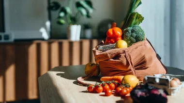 A cloth bag filled with fresh vegetables and fruits, including broccoli, tomatoes, and lemons, sits on a wooden table.