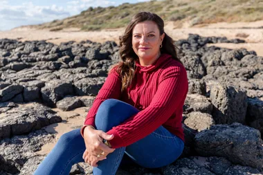 Woman in a red sweater and jeans sitting on rocky beach with greenery and cloudy sky in the background.