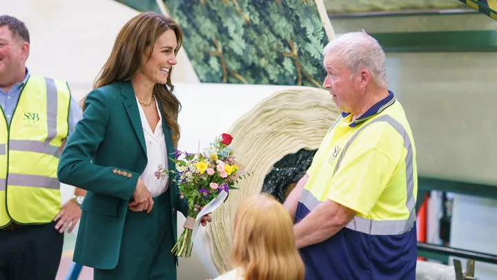 A woman in a green suit holding flowers speaks with a factory worker in a yellow vest; background shows rolled materials.