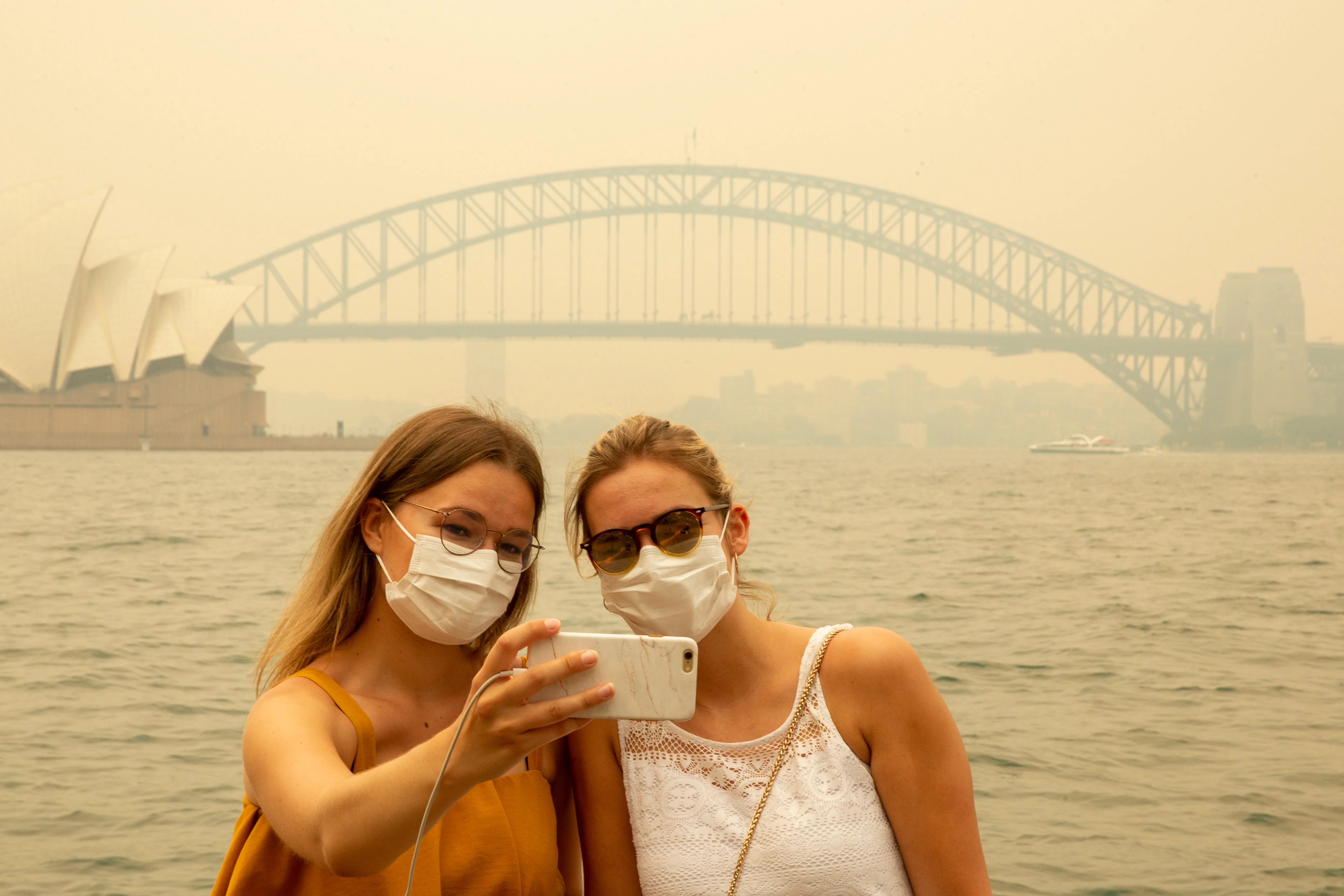 SYDNEY, AUSTRALIA - DECEMBER 19: German tourists Julia Wasmiller (L) and Jessica Pryor take a selfie at Mrs Macquarie's chair, wearing face masks due to heavy smoke on December 19, 2019 in Sydney, Australia. NSW Premier Gladys Berejiklian has declared a state of emergency for the next seven days with ongoing dangerous fire conditions and almost 100 bushfires burning across the state. It's the second state of emergency declared in NSW since the start of the bushfire season. (Photo by Jenny Evans/Getty Images)