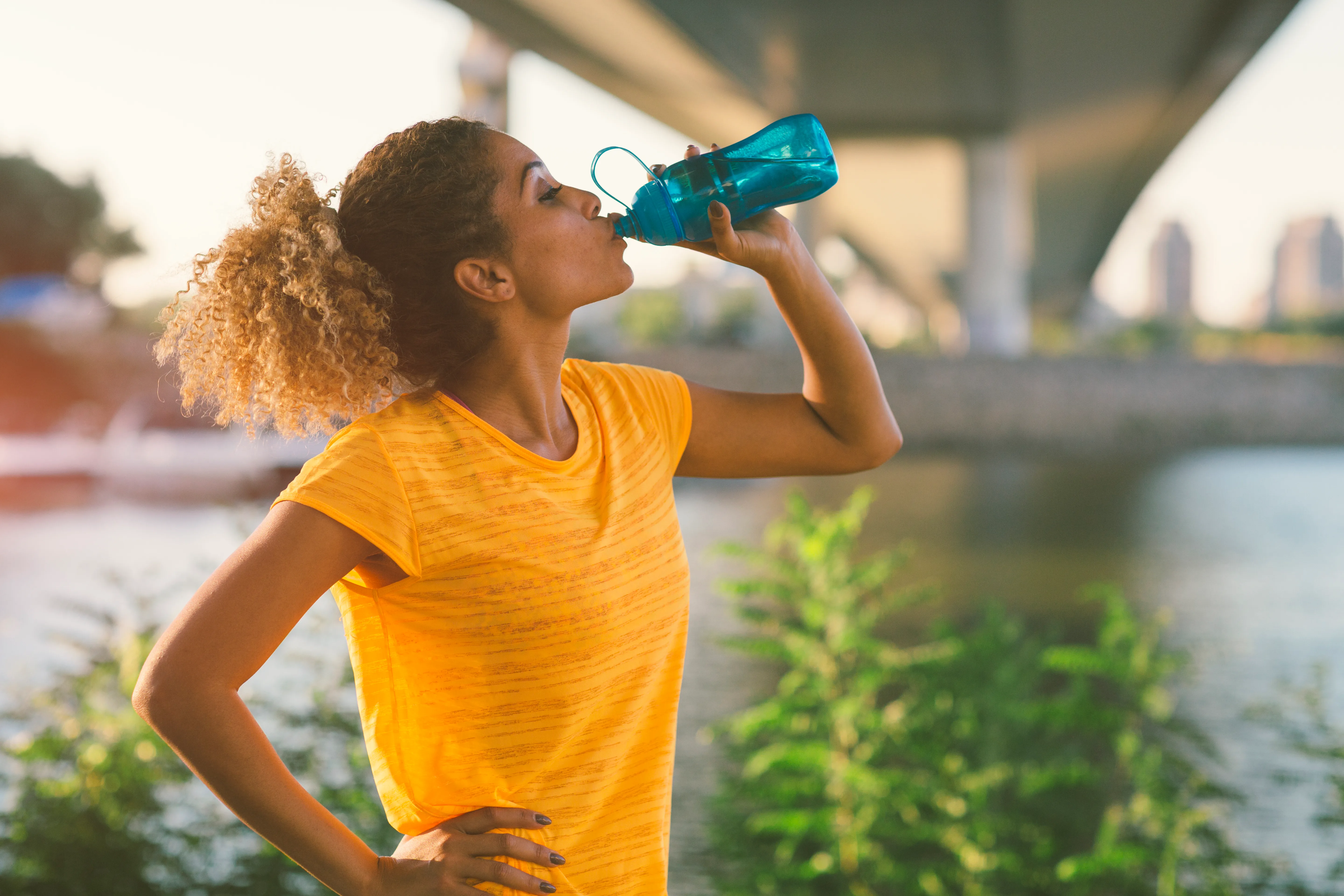 Latina Runner running in the city near river. Standing under the bridge, making break to drink some water. Cityscape in background.