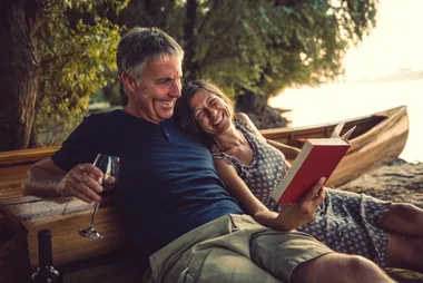 Couple relaxing by a river, woman reading a book, and man holding a glass while sitting on a canoe.