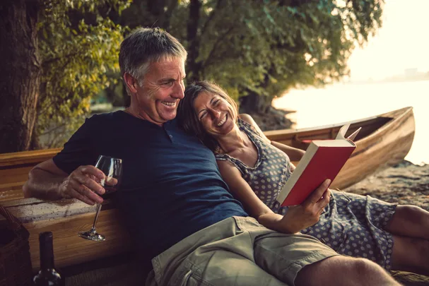 Couple relaxing by a river, woman reading a book, and man holding a glass while sitting on a canoe.