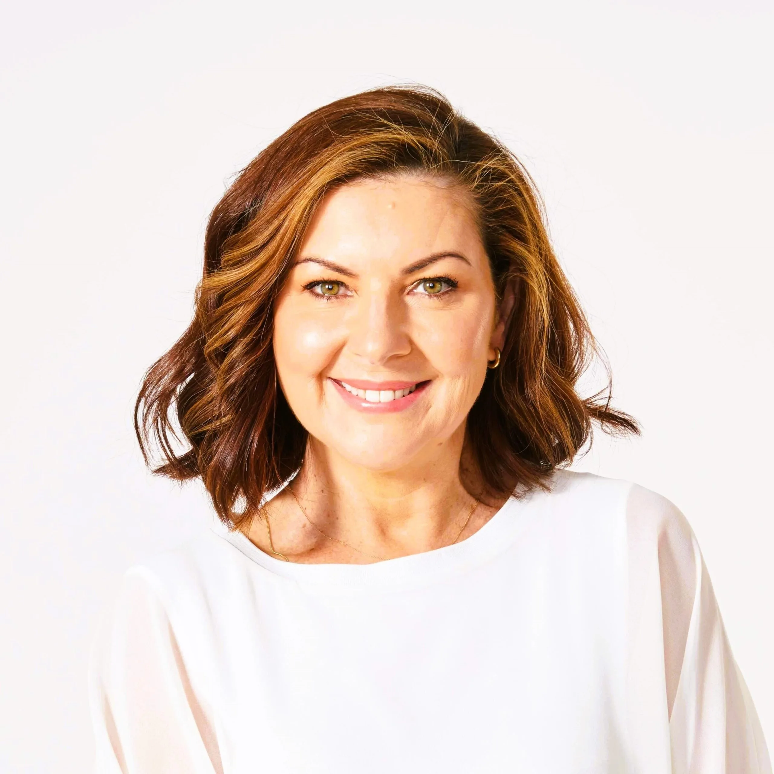A woman with shoulder-length brown hair smiling, wearing a white top, against a light background.